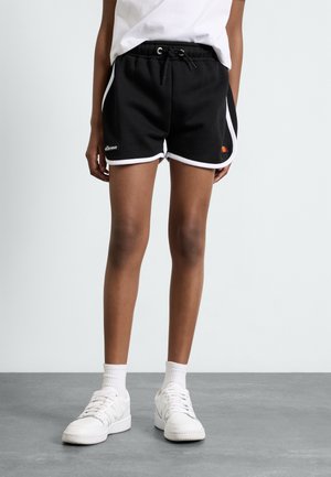 Person trägt schwarze Sportshorts mit weißem Saum, weiße Socken und weiße Turnschuhe, steht auf grauem Boden vor hellem, einfarbigem Hintergrund.