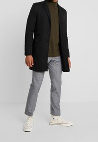 Zwarte jas met twee knopen en zijzakken, olijfgroene coltrui, grijs geruite broek en witte hoge sneakers.