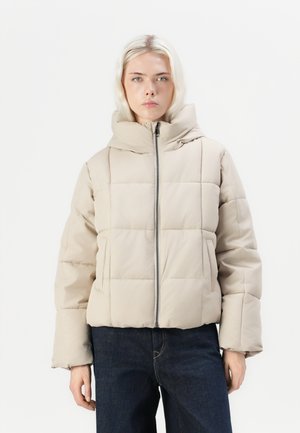 Veste beigne matelassée avec fermeture éclair sur le devant et col montant ; dotée d'un motif matelassé et de poignets élastiques ; portée sur un jean bleu foncé.