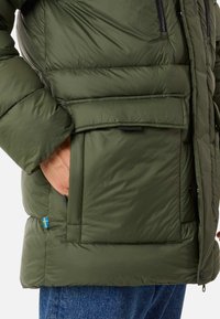 Olive green puffer jacket with a quilted texture, featuring a front pocket, zip closure, and a small Swedish flag detail on the side.