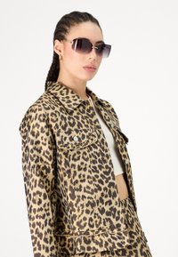 Denim jakna s leopardskim printom, ovratnikom, prednjim žepovima in srebrnimi gumbi. Model nosi temne sončne očala; ozadje je belo.