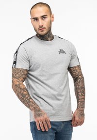 Tricou gri cu mânecă scurtă și guler rotund, având dungi negre pe laterale și un logo mic pe piept. Material din bumbac cu o textură netedă.