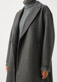 Grå ullrock med bred krage, sänkt axel och sidofickor, lager-på-lager över en texturerad ljusgrå polo-tröja.