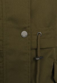 Veste vert olive en coton texturé avec fermeture à pression métallique, cordon de serrage et poches doubles. Présente le logo VERO MODA sur la pression.