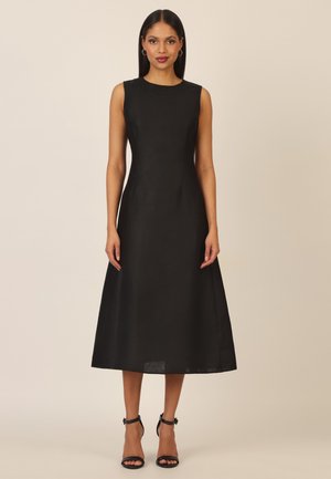 Woman standing wearing a sleeveless black midi dress and black ankle-strap high heels against a plain beige background.