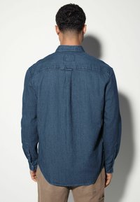 Donkerblauwe denimshirt met lange mouwen, knoopmanchetten en een ruginzet. Heeft een klassieke kraag en een label op de hals.