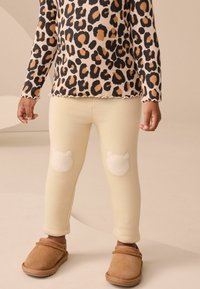 Camicia a maniche lunghe in stampa leopardata beije, abbinata a leggings color crema con toppe sulle ginocchia a forma di orso chiaro e slippers marroni.