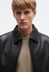 Fekete bőrdzseki ing gallérral és elöl cipzárral, világos bézs, texturált pulóver fölött viselve. Semleges háttér.