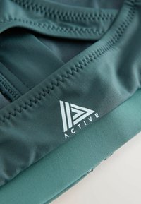 Træningstøjsstof i dyb grøn med en glat tekstur. Har en kontrasterende logo i hvid samt flatlock-syninger langs kanterne.