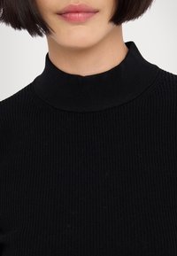 Gros plan d'une personne portant un pull noir côtelé à col montant, avec des cheveux courts et foncés visibles au niveau de la mâchoire.