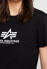 Camiseta de algodón negra con un logo blanco que presenta un diseño de avión y el texto "ALPHA INDUSTRIES KNOXVILLE, TENNESSEE, U.S.A."