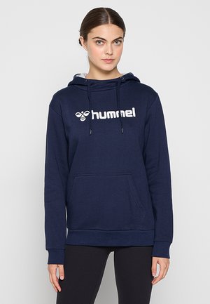 Kvinde med mørkt hår sat tilbage iført en marineblå hættetrøje med "hummel" logo og sorte bukser står mod en ensfarvet lys baggrund.