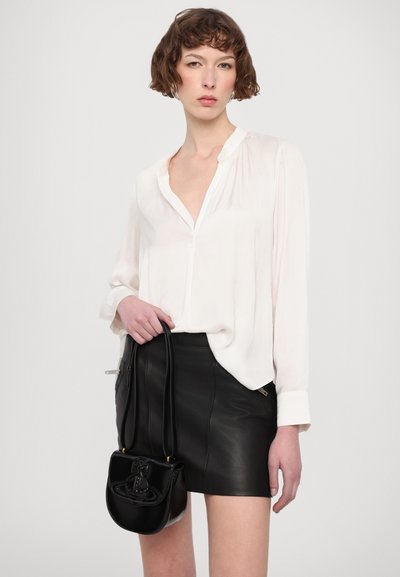 Femme aux cheveux courts bruns portant un chemisier blanc et une mini-jupe en cuir noire, tenant un petit sac à main noir par ses anses.