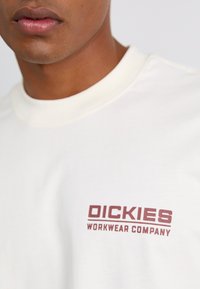 T-shirt in cotone crema con stampa maroon "DICKIES WORKWEAR COMPANY" sul petto. Collo rotondo con una texture liscia.