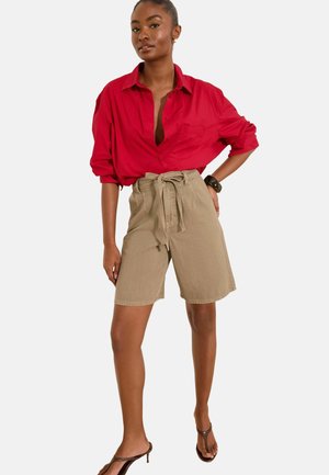 Kvinde iført en løs rød skjorte med knapper, beige højtaljede shorts med bindebånd, mørkt armbånd og brune hælede sandaler, poserer med den ene hånd på hoften.
