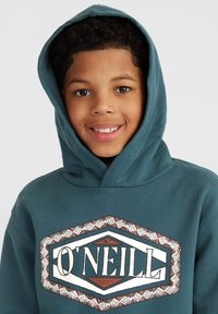 Felpa color teal con un patch grafico frontale che presenta il nome del marchio "O'NEILL". Tessuto morbido, cappuccio con cordini e motivo decorativo attorno al patch.