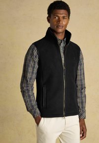 Melnas vilnas veste ar augstu apkakli, ar rāvējslēdzēju un sānu kabatām, valkāta virs raibā pogājamā krekla ar daudzkrāsainiem kvadrātiem.