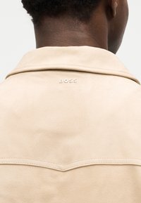 Άτομο με σκουρόχρωση δέρματος που φοράει μπεζ μπουφάν με γιακά, με τη λέξη "BOSS" κεντημένη στο πάνω μέρος της πλάτης.