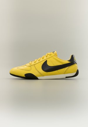 Geltoni žemi sportbačiai su juodu Nike logotipu ir akcentais, balta pado dalimi bei raištelių dizainu ant neutralaus fono.