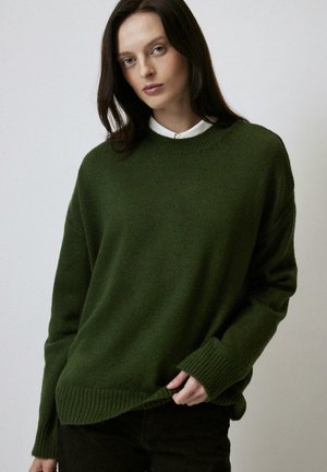 Dark green knit sweater with a round neck, ribbed cuffs, and hem. Features a relaxed fit and subtle texture. Worn over a white collared shirt.