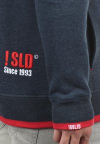 Sudadera navy con acentos rojos, puños de canalé y texto bordado "! SLD Desde 1993." Cuenta con un bolsillo lateral y tela texturizada.