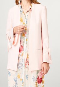 Kobieta w jasnoróżowej marynarce na kwiecistej sukience z czerwonymi, żółtymi i niebieskimi kwiatami, trzymająca klapę marynarki jedną ręką.