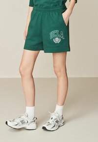 Zaļi kokvilnas šorti ar elastīgu jostasvietu, uz kuriem redzams balts "UCLA" logo un emblēma. Savienoti ar baltām sporta kurpēm un potīšu zeķēm.