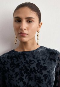 Vrouw met naar achteren gekamd haar, draagt lange gouden hangende oorbellen en een zwarte fluwelen top met bloemmotief, tegen een effen witte achtergrond.