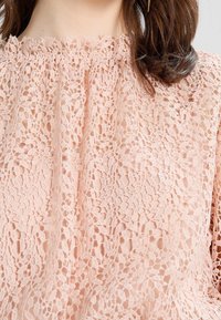 Top in pizzo di un morbido rosa con un motivo floreale a rilievo, scollatura arricciata e vestibilità ampia e leggera.
