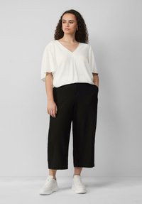 Witte V-halsblouse met korte mouwen, zwarte brede broek en witte sneakers. Krullend haar, staand tegen een grijze achtergrond.