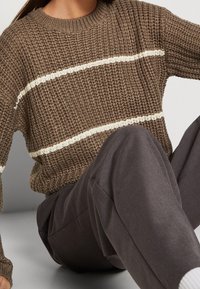 Brązowy sweter z dzianiny o strukturalnym wykończeniu i białymi poziomymi paskami, w połączeniu z ciemnoszarymi spodniami o luźnym kroju.