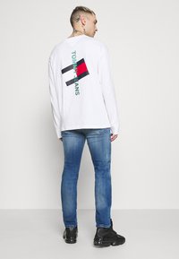 White long-sleeve T-shirt featuring a graphic design with black, red, and blue elements, paired with light blue denim jeans and black sneakers.