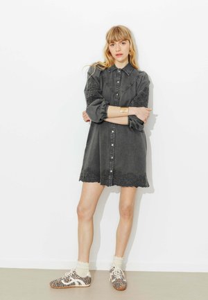 Femme debout les bras croisés, portant une robe boutonnée gris foncé avec broderies, des baskets à imprimé léopard et des chaussettes crème.