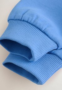 Manšetni rokavi svetlo modrega puloverja z rebrasto teksturo. Tkanina je videti mehka, pri šivih rahlo raztegljiva z gladkim zaključkom.
