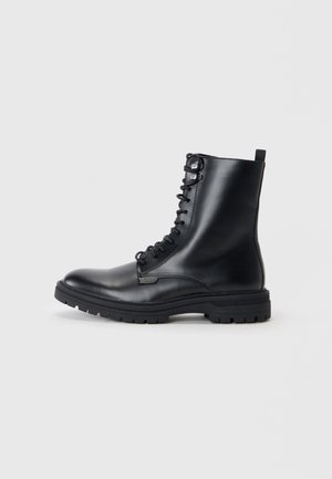 Botas de combate de cuero negro con cordones, punta redonda, acabado texturizado y suela de goma gruesa con surcos profundos. Asa en la parte posterior.