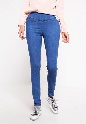 Žena v modrých úzkých džínách s V-znakem v pase a stříbrnými metalickými šněrovacími botami, stojící na bílém pozadí.