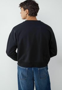 Czarna bluza z okrągłym dekoltem i ściągaczami, wykonana z miękkiego materiału, zestawiona z niebieskimi dżinsami. Prosty, luźny krój.