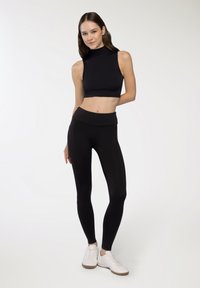 Czarny bezrękawnikowy crop top z wysokim kołnierzem, zestawiony z czarnymi legginsami o wysokim stanie. Model nosi białe sneakersy. Gładki, elastyczny materiał.
