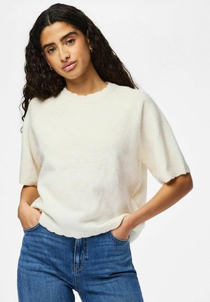 Mujer con cabello rizado que lleva un suéter blanco de manga corta y jeans azules, con las manos en los bolsillos, de pie contra un fondo liso.