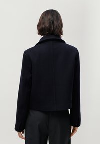 Jachetă dintr-un amestec de lână, de culoare bleumarin, cu guler clasic, croială scurtă, umeri ușor structurați și textură netedă. Spate curat, fără accesorii.