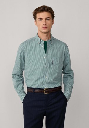 Hombre con camisa de cuadros verdes y blancos de botones, pantalones de raya diplomática azul oscuro y cinturón trenzado marrón, con las manos en los bolsillos, fondo neutro.