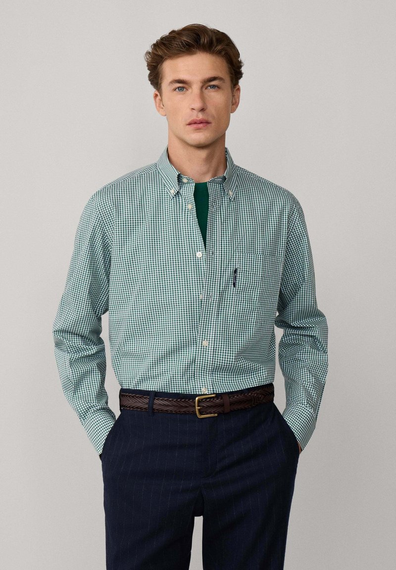 Hombre con camisa de cuadros verdes y blancos de botones, pantalones de raya diplomática azul oscuro y cinturón trenzado marrón, con las manos en los bolsillos, fondo neutro.