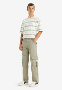 Mlad moški stoji, oblečen v belo in zeleno prugasto majico s kratkimi rokavi, svetlo zelenih hlačah s cargo žepi in belih supergah s črnimi detajli.