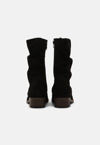 Bottines noires en suède hautes jusqu'aux genoux avec un design ample, des zips sur les côtés et un talon en bois empilé. Texture lisse et accents minimalistes.