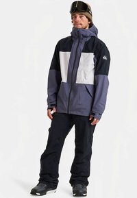 Άνδρας που φοράει μαύρο και γκρι σακάκι σκι, μαύρο παντελόνι χιονιού, μαύρες μπότες χιονιού, πράσινο σκουφί και μαύρες γυαλιά σκι στο κεφάλι.