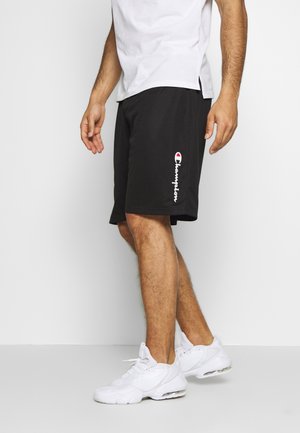 Homme portant un short noir Champion, des chaussures de sport blanches et un T-shirt blanc, debout sur un fond blanc uni.