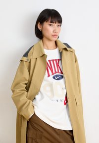 Veste oversize kaki avec un col gris, superposée à un t-shirt blanc arborant des graphiques rouges et bleus de Nike, accompagnée de pantalons cargo marron.