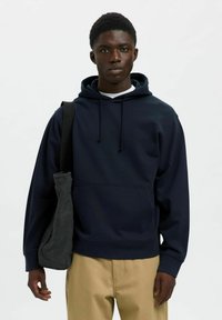 Sudadera azul marino con capucha ajustable, bolsillo frontal y puños de canalé. Combinada con pantalones beige claros y un bolso oscuro de hombro.