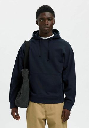 Sudadera azul marino con capucha ajustable, bolsillo frontal y puños de canalé. Combinada con pantalones beige claros y un bolso oscuro de hombro.