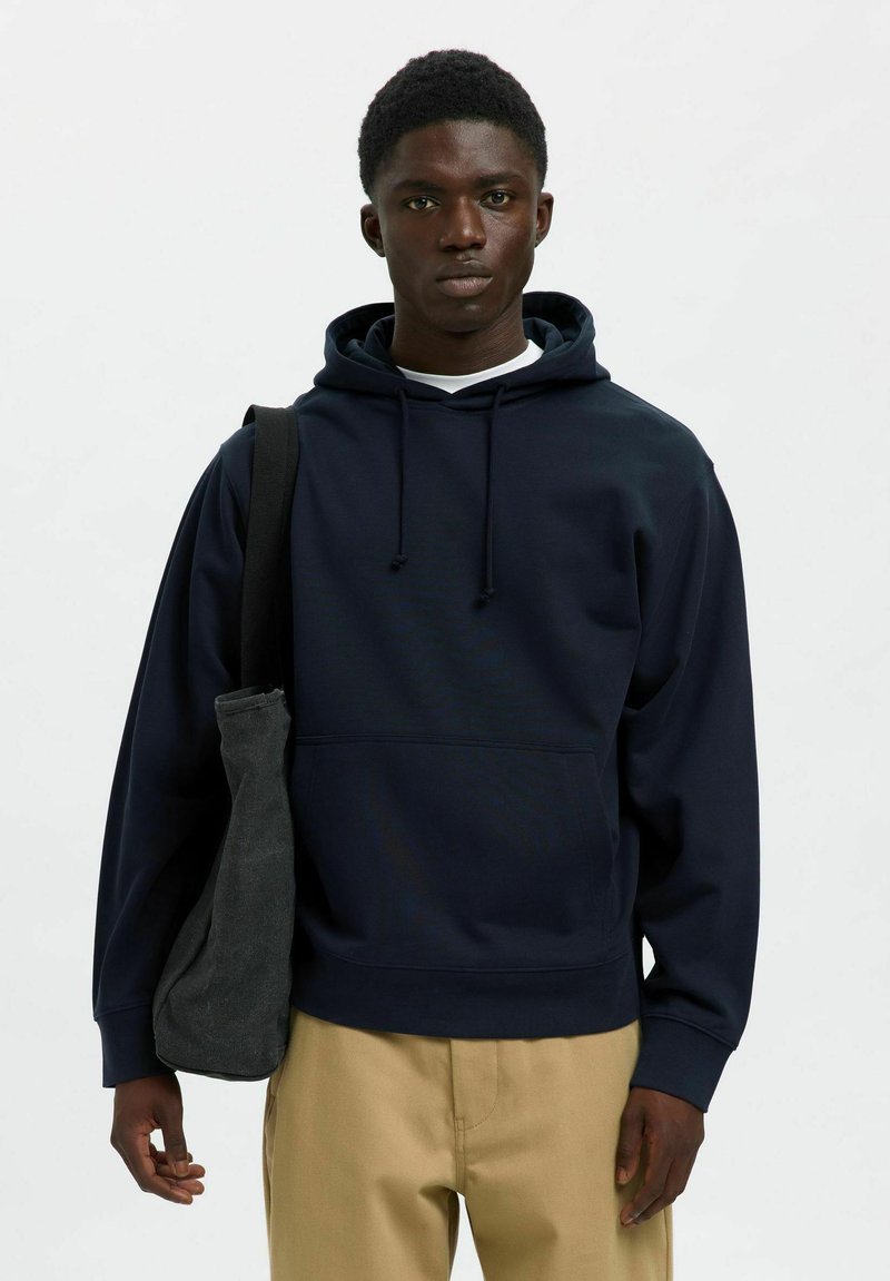Sudadera azul marino con capucha ajustable, bolsillo frontal y puños de canalé. Combinada con pantalones beige claros y un bolso oscuro de hombro.